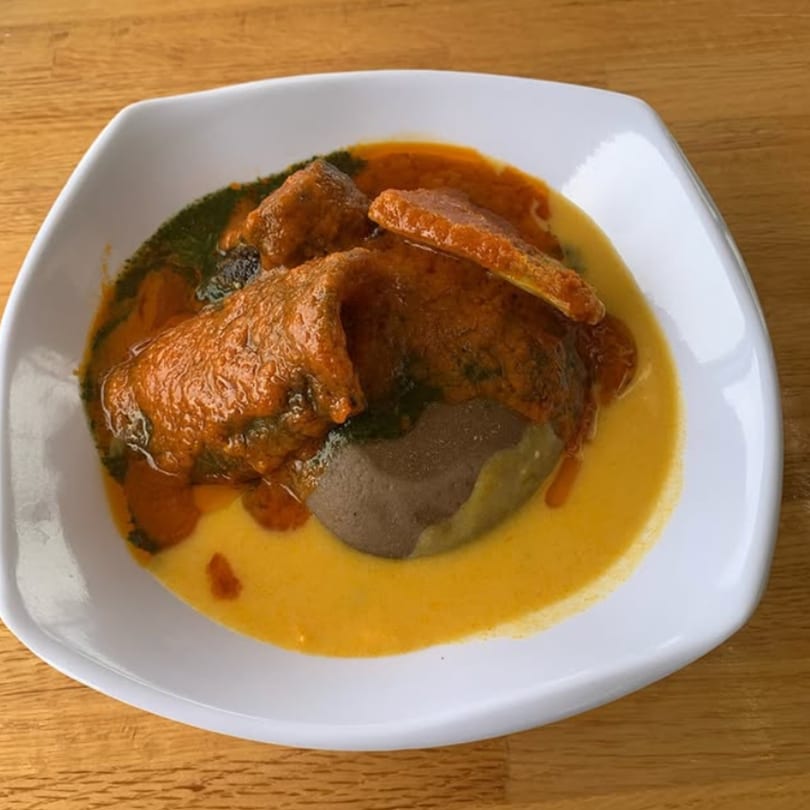 A white square plate holds a serving of African stew with fufu, surrounded by orange and yellow sauces.