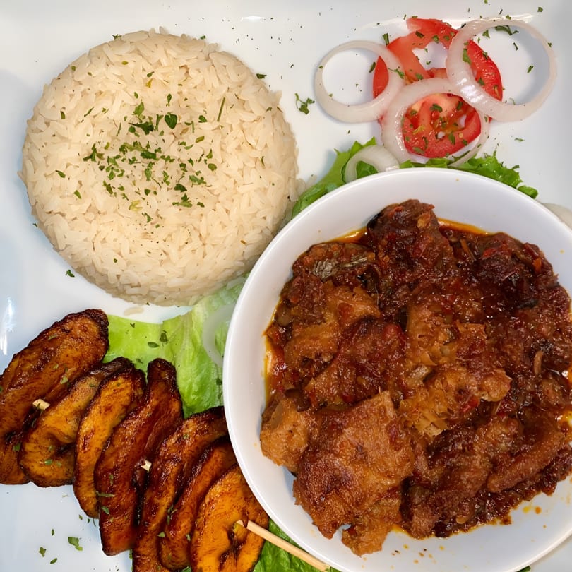 A plate of African food with white rice, stewed meat, fried plantains, and a side salad.