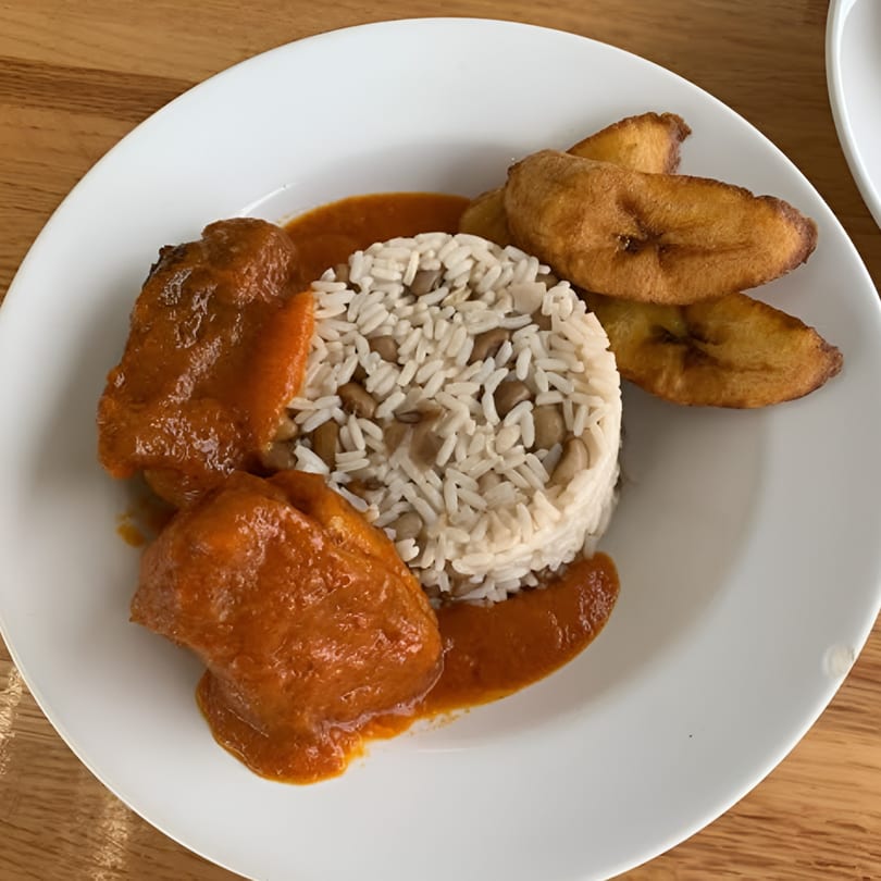 African dish with chicken in red sauce, rice and beans, and fried plantains on a white plate.